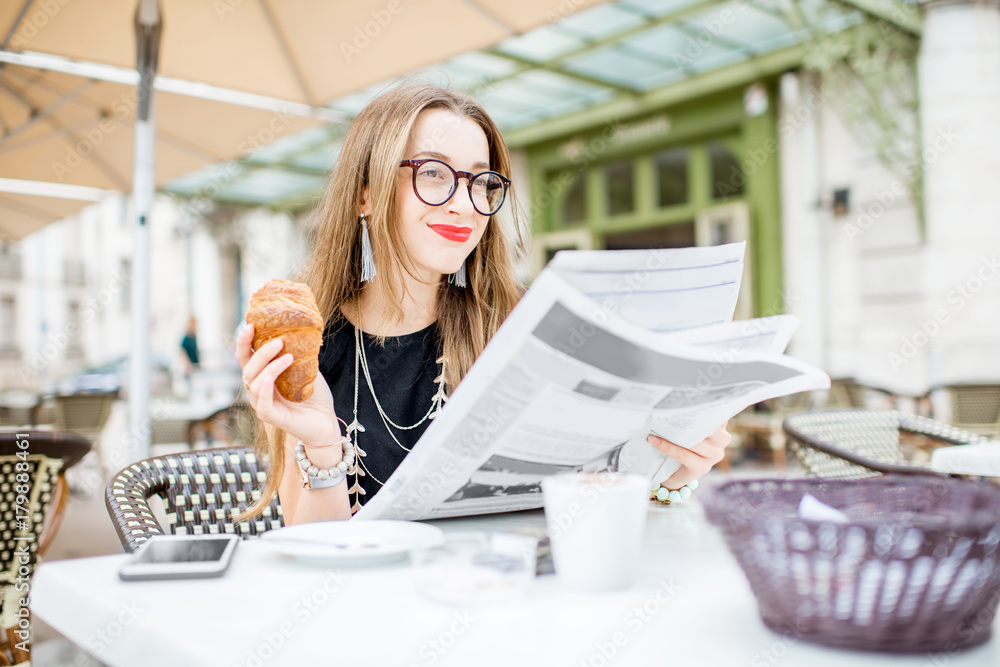 Young woman reading newspaper during a breakfast with croissant sitting outdoors at the typical fren