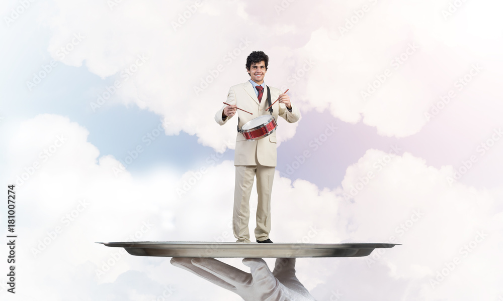 Young businessman on metal tray playing drums against blue sky background