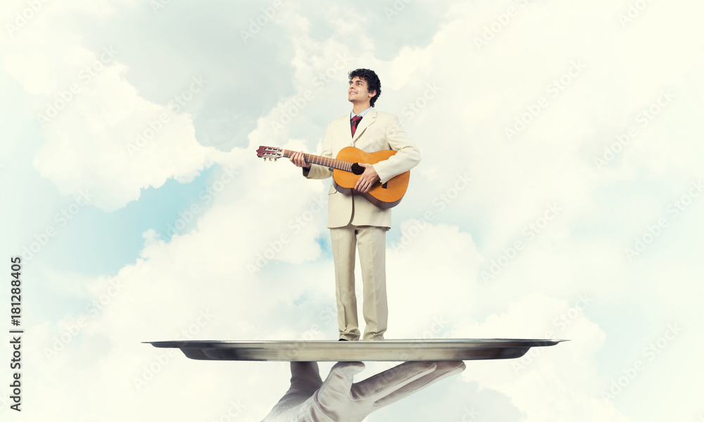 Businessman on metal tray playing acoustic guitar against blue sky background