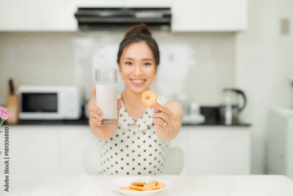 年轻的亚洲女人在厨房里拿着牛奶杯咬饼干