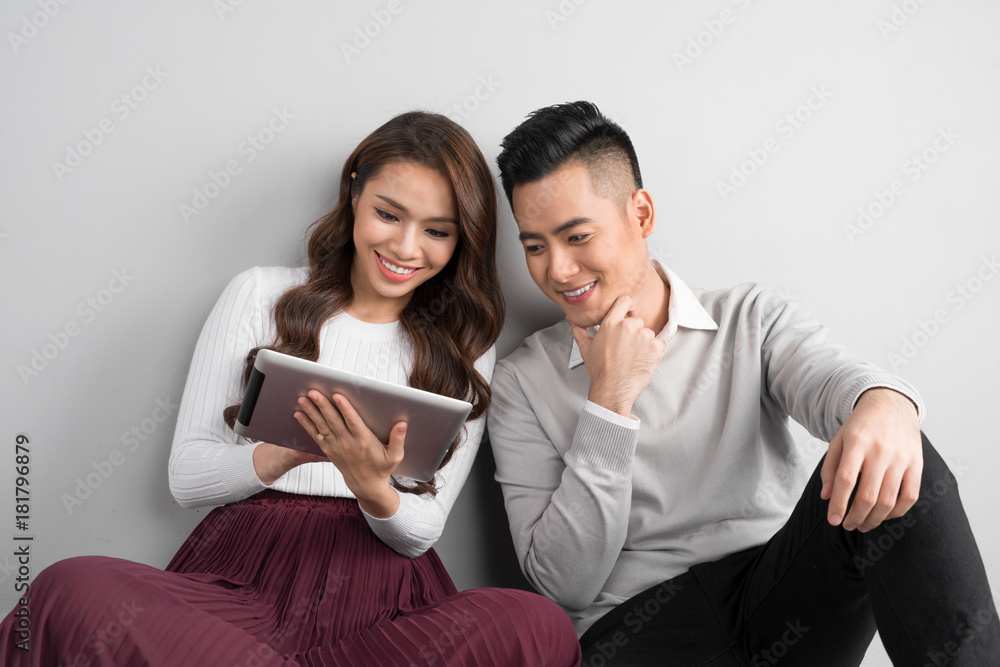 Pretty smiling asian couple in love together sitting on floor enjoying using digital tablet