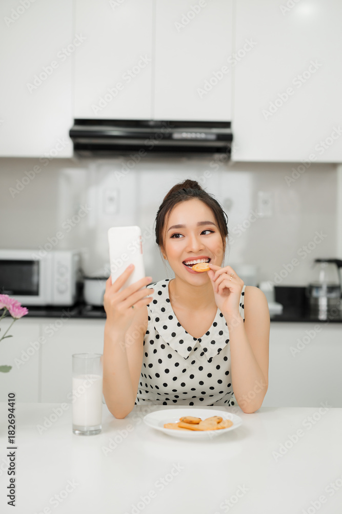 年轻微笑的女人吃着饼干，微笑着，使用智能手机。健康的早餐。