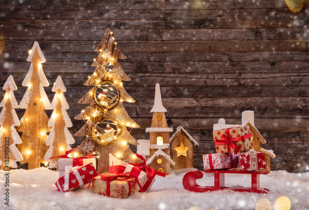 Holidays background with illuminated Christmas tree, sledge with gifts and wooden village.