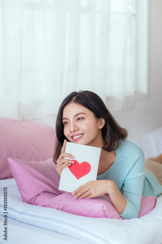 亚洲女孩在家躺在床上，开心地拿着红色心形的贺卡。