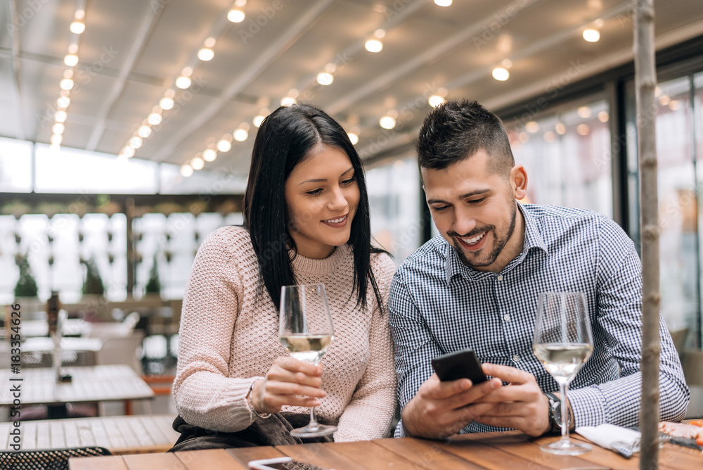 Portrait of happy young man and woman sitting together at a restaurant and looking at a mobile phone