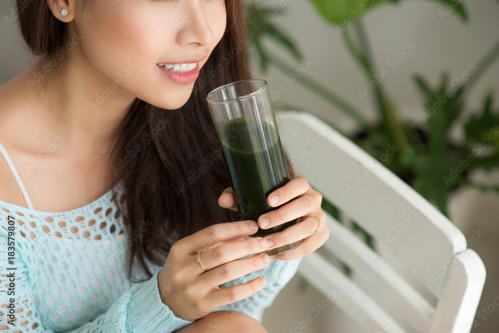 Smiling young asian woman drinking green fresh vegetable juice or smoothie from glass at home