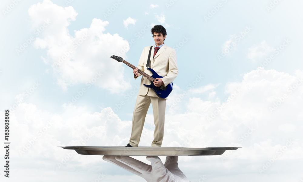 Businessman on metal tray playing electric guitar against blue sky background