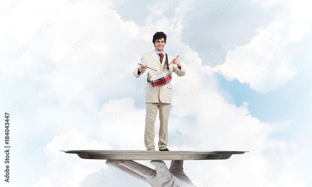 Young businessman on metal tray playing drums against blue sky background