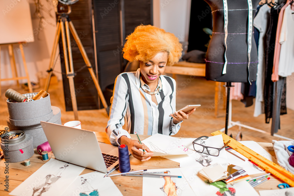 Young african fashion designer working with clothes drawings and laptop sitting at the studio interi