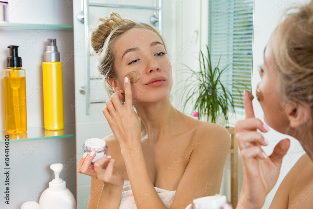 Young beautiful girl applying facial scrub mask on skin. Looking in the mirror in bathroom, Wrapped 