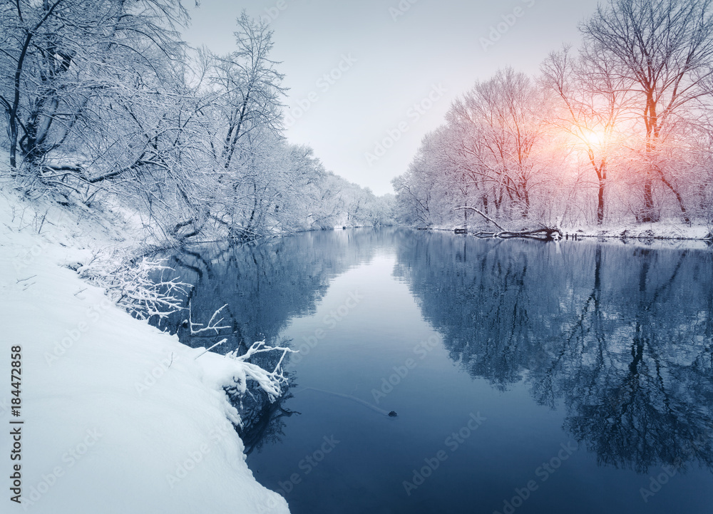 日落时河边的冬季森林。白雪皑皑的树木点缀着五颜六色的景观，冰冻的河流倒影着