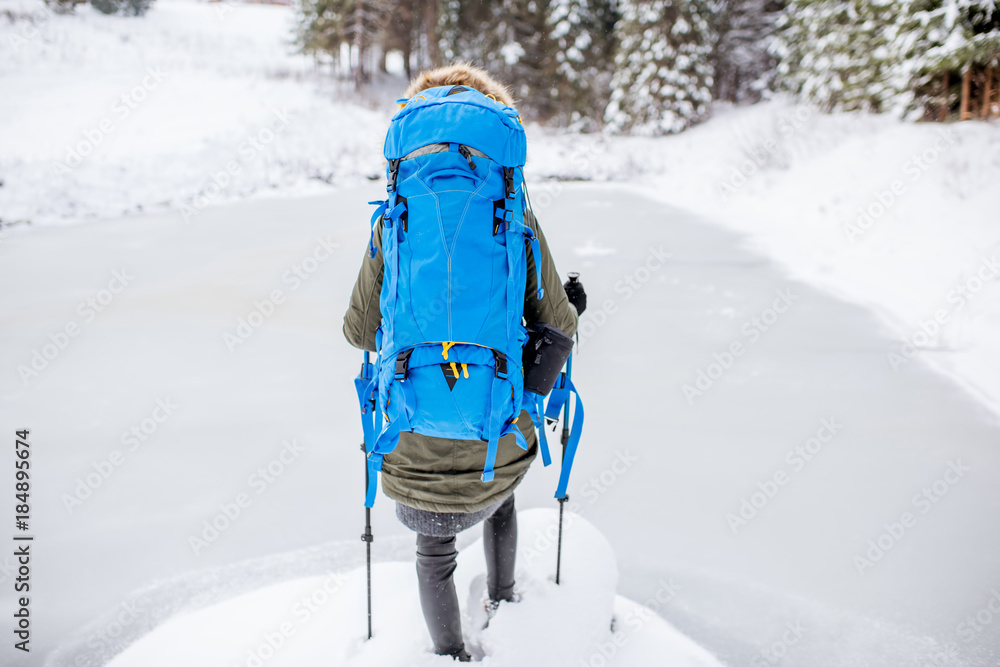 Woman in winter clothes with backpack and tracking sticks standing near the frozen lake during the w