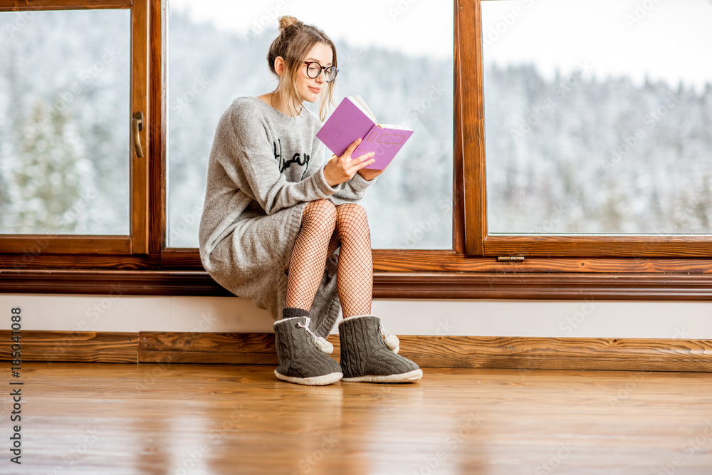 Young woman in sweater reading book near the window at the cozy wooden mountain house with beautiful