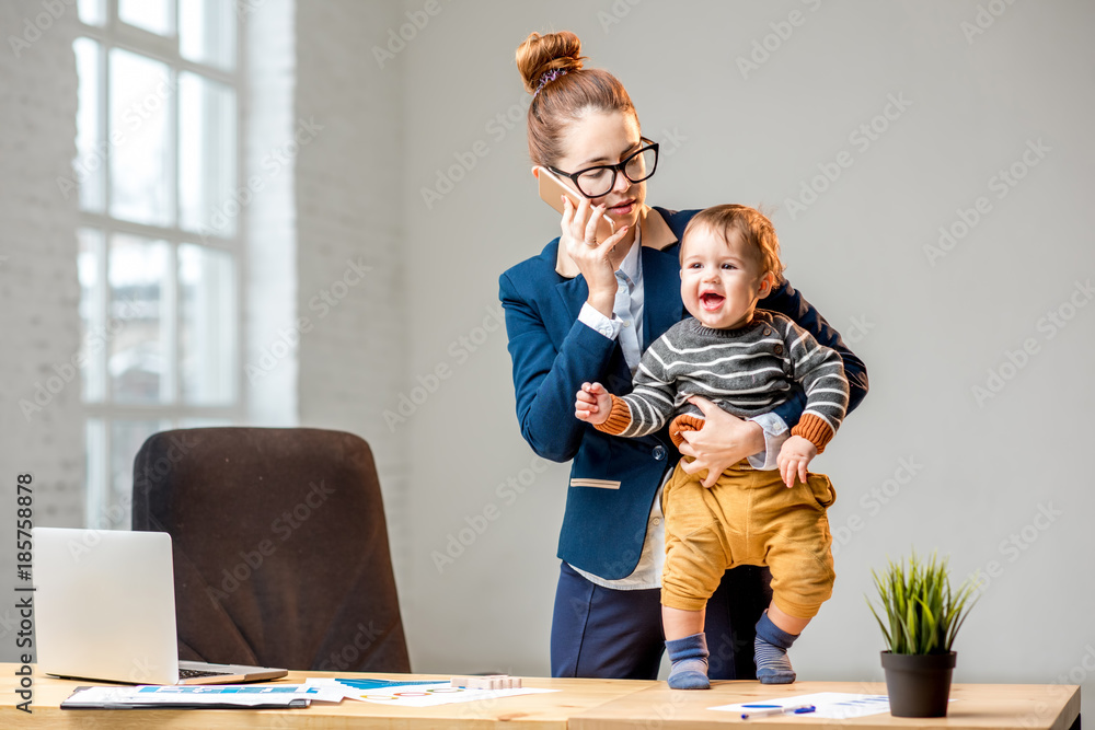 Young multitasking businessmam dressed in the suit talking phone standing with her baby son at the o