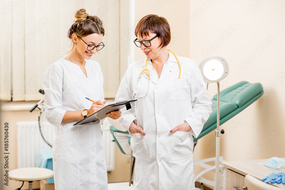 Senior doctor talking with young woman assistant standing in the gynecological office with chair and