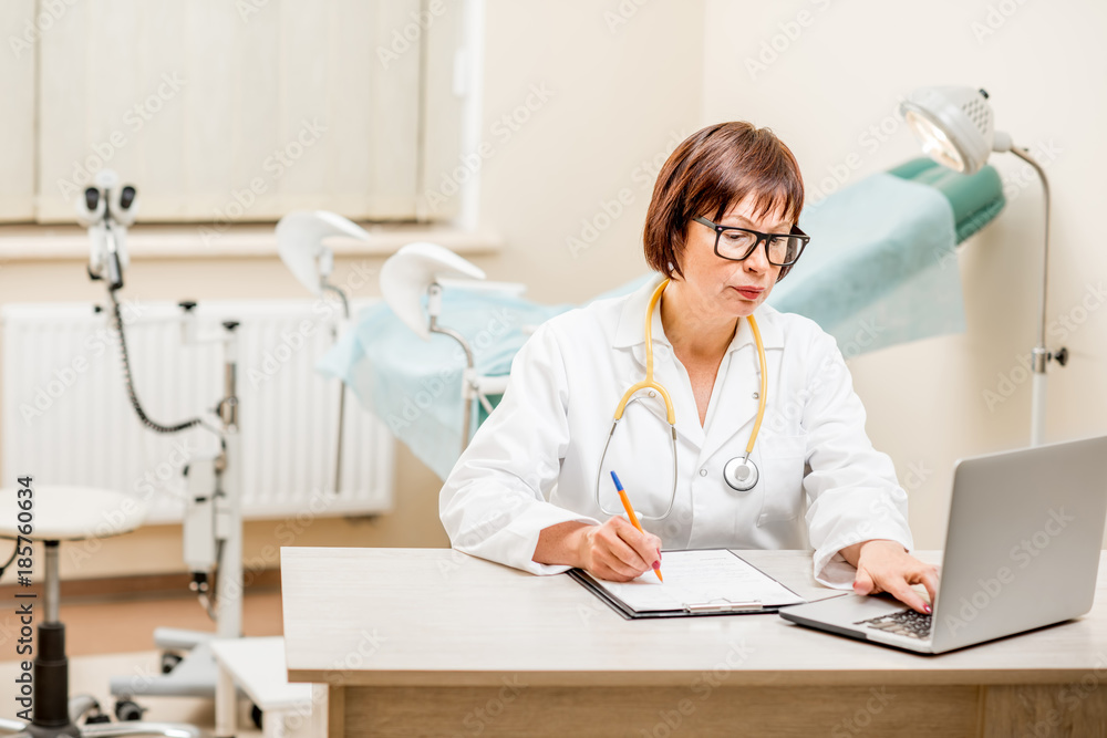 Seinor doctor working with laptop and documents sitting in the gynecological office with gynecologic