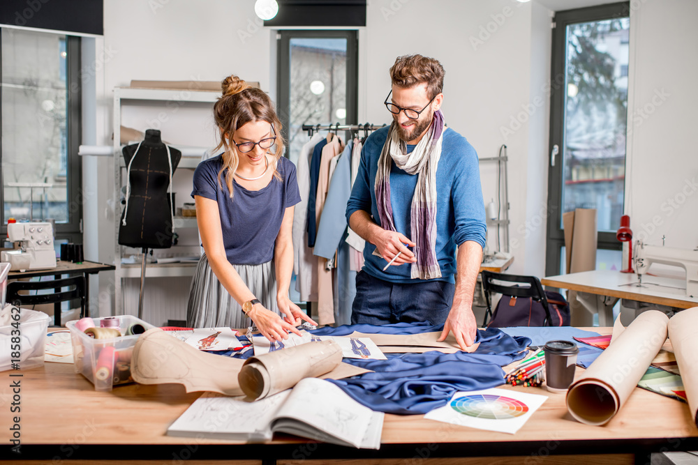 Couple of fashion designers working with fabric and clothing sketches at the studio full of tailorin