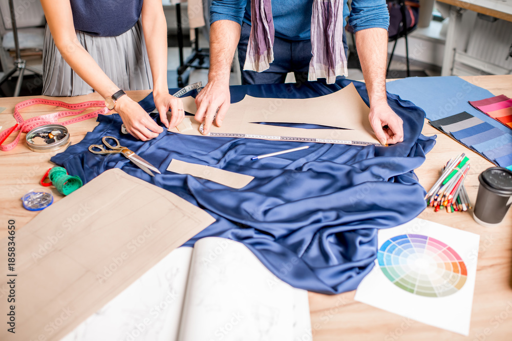 Cutting blue fabric on the table full of tailoring tools. Close-up view on the hands and fabric with