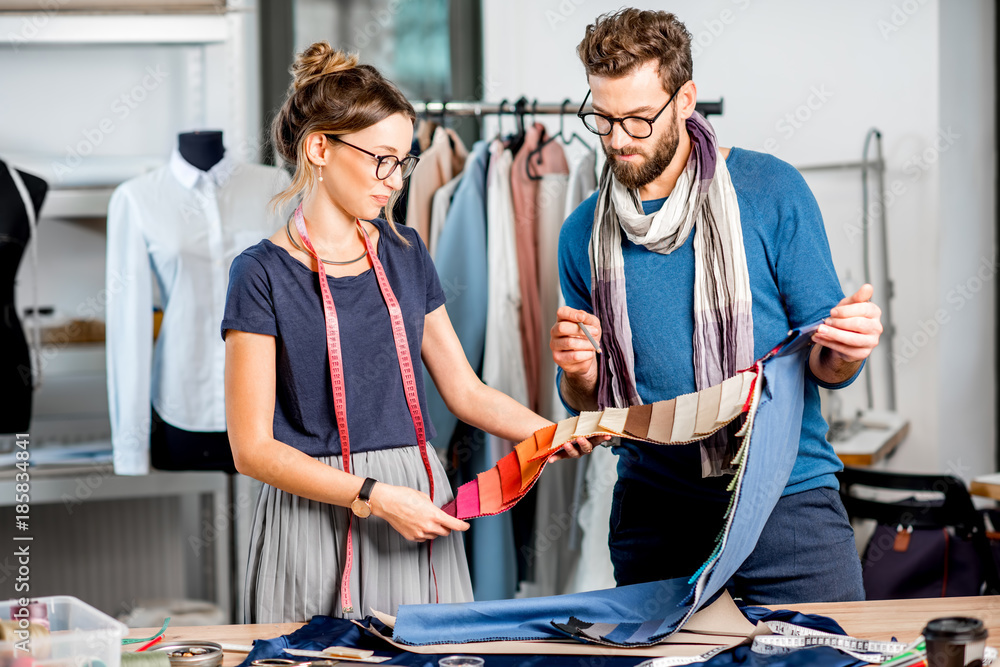 Couple of fashion designers choosing fabric standing at the studio full of tailoring tools and equip