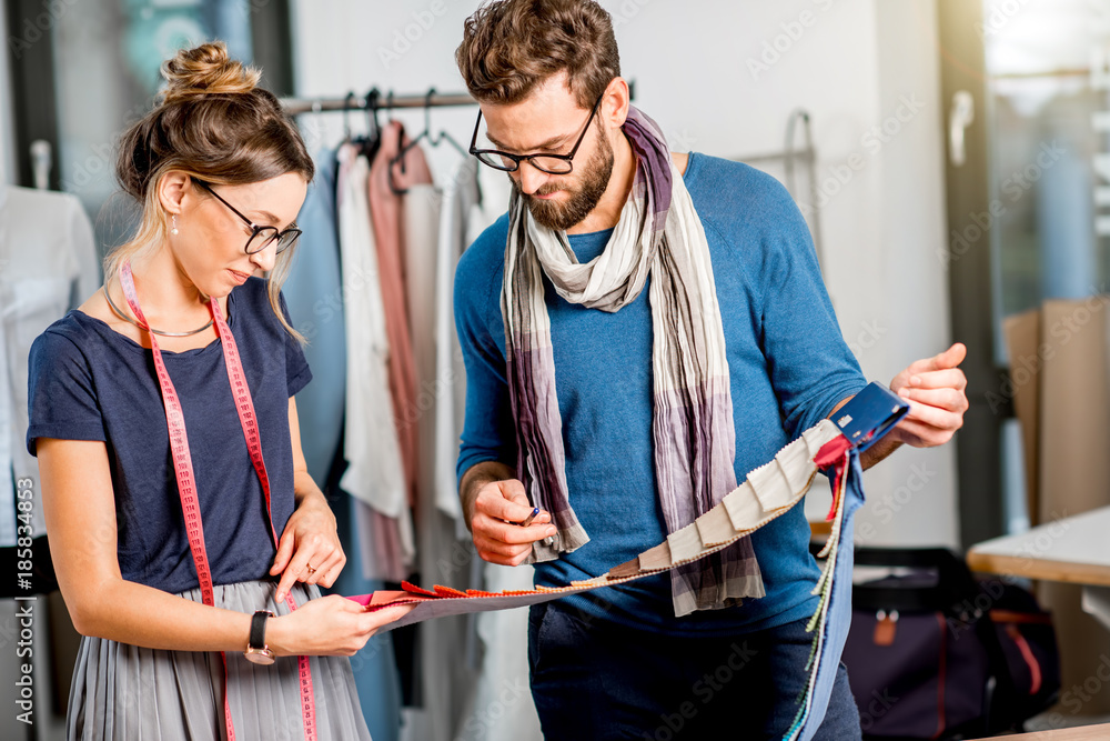 Couple of fashion designers choosing fabric standing at the studio full of tailoring tools and equip