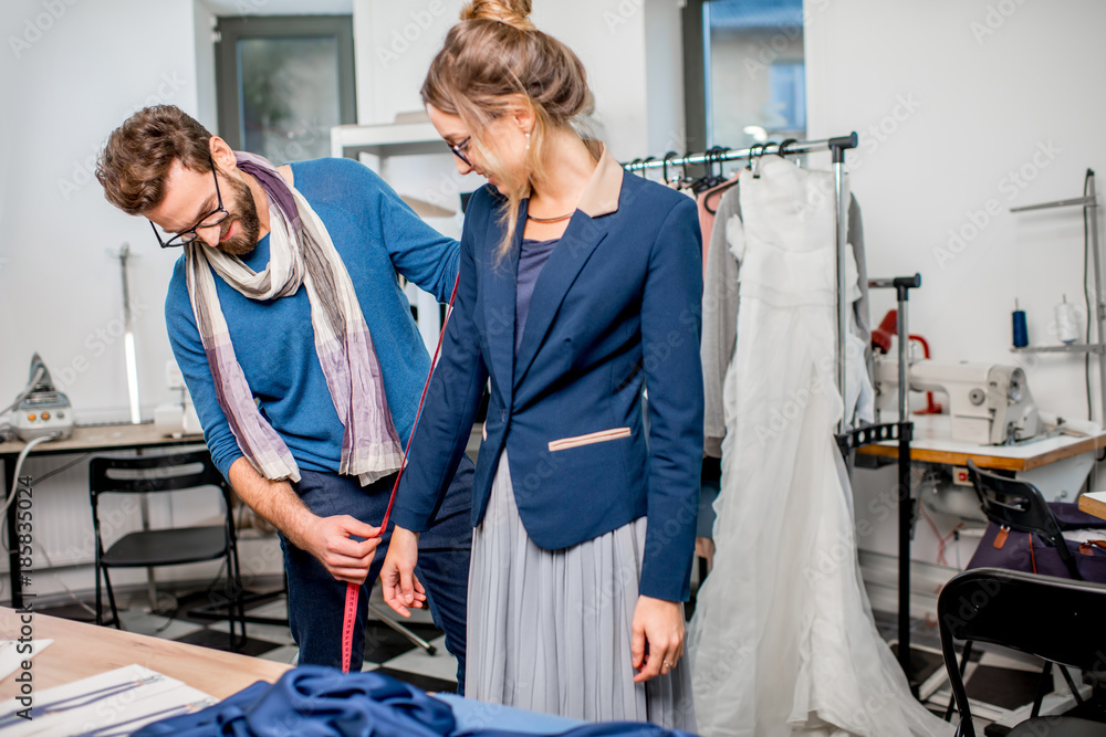 Handsome tailor measuring jacket on the woman client standing at the studio full of tailoring tools 