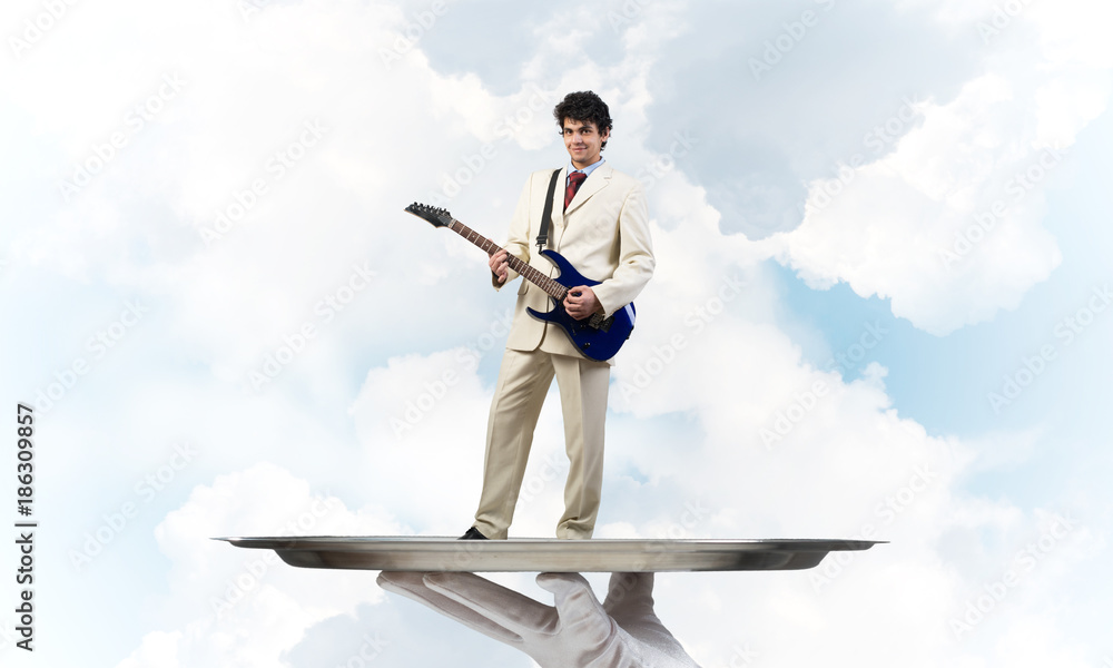 Businessman on metal tray playing electric guitar against blue sky background