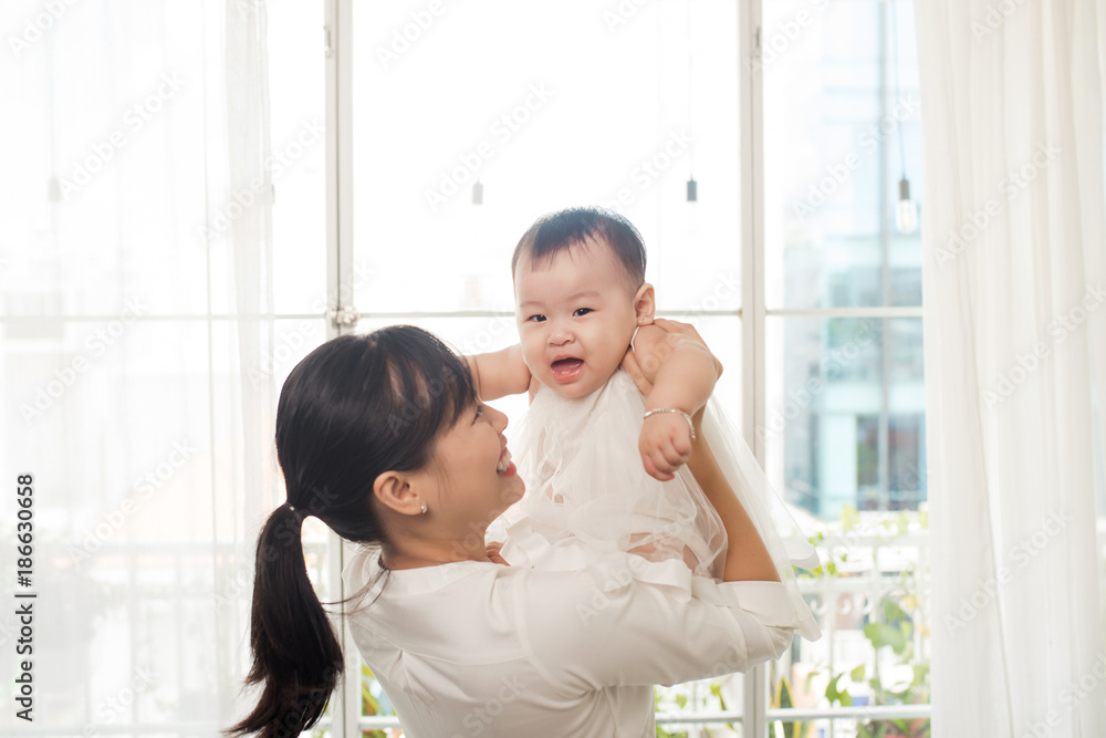 快乐的亚洲母亲和婴儿在窗户附近的白色房间里一起玩耍的肖像