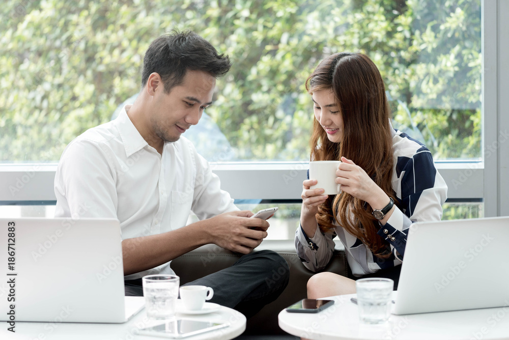 Two asian coworkers use laptop and smartphone work together having coffee in afternoon