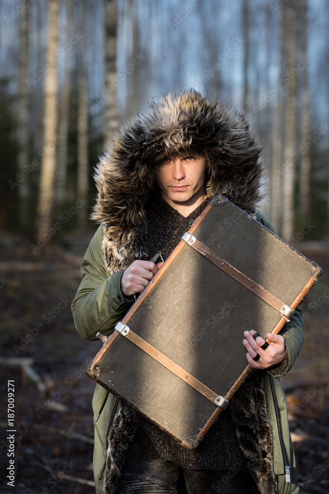 Young man in a green winter parka in a forest with a suitcase.