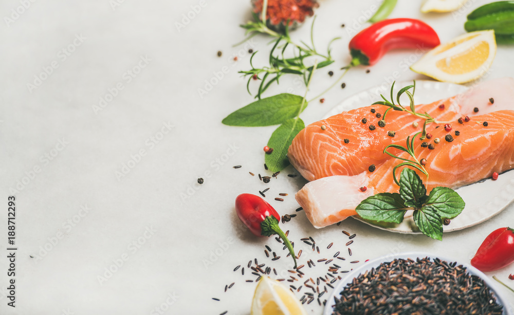 Raw salmon fish fillet steaks with vegetables, greens, rice, spices and lemon over light grey marble