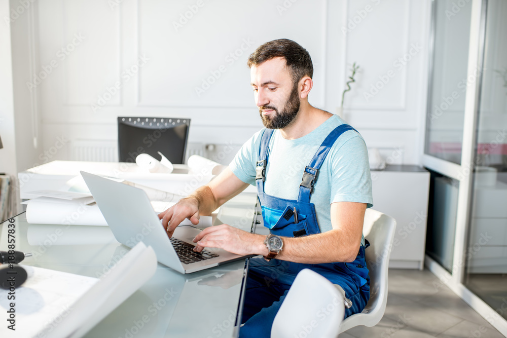 Handsome repairman or foreman in uniform working with laptop and architectural drawings at the offic