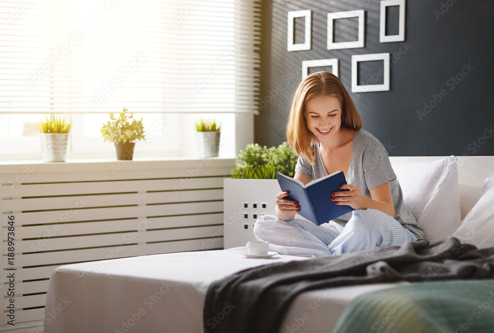 Happy young woman reads  book and drinks coffee in bed