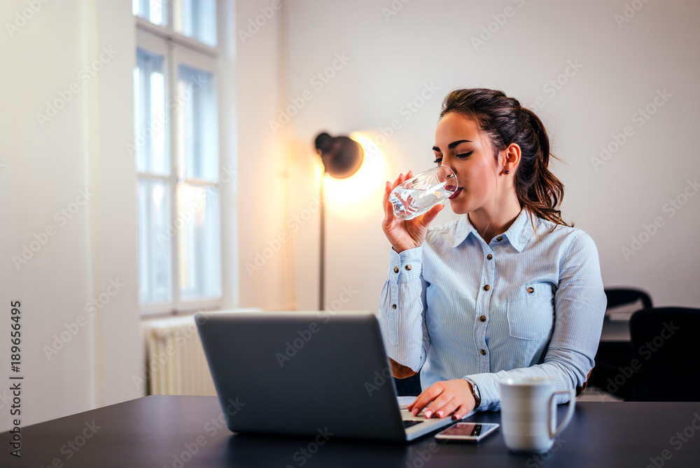 微笑的女商人在办公室的办公桌旁喝着一杯水。