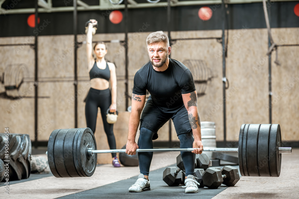 Handsome athletic man in black sports wear lifting up a heavy burbell with woman training on the bac