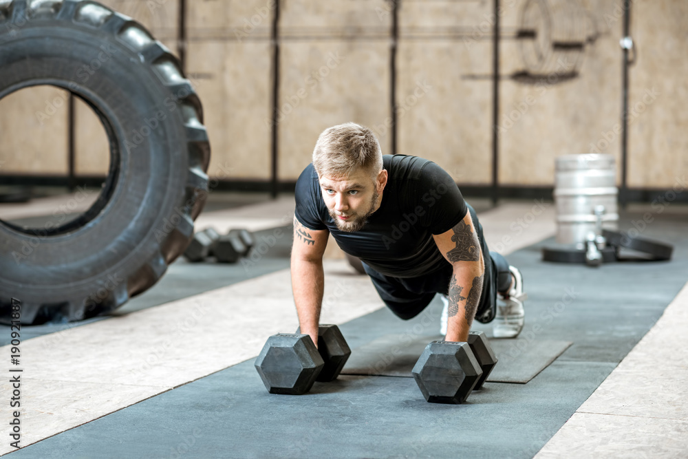 Athletic man in black sportswear with tattoos pushing ups on the dumbbells in the gym