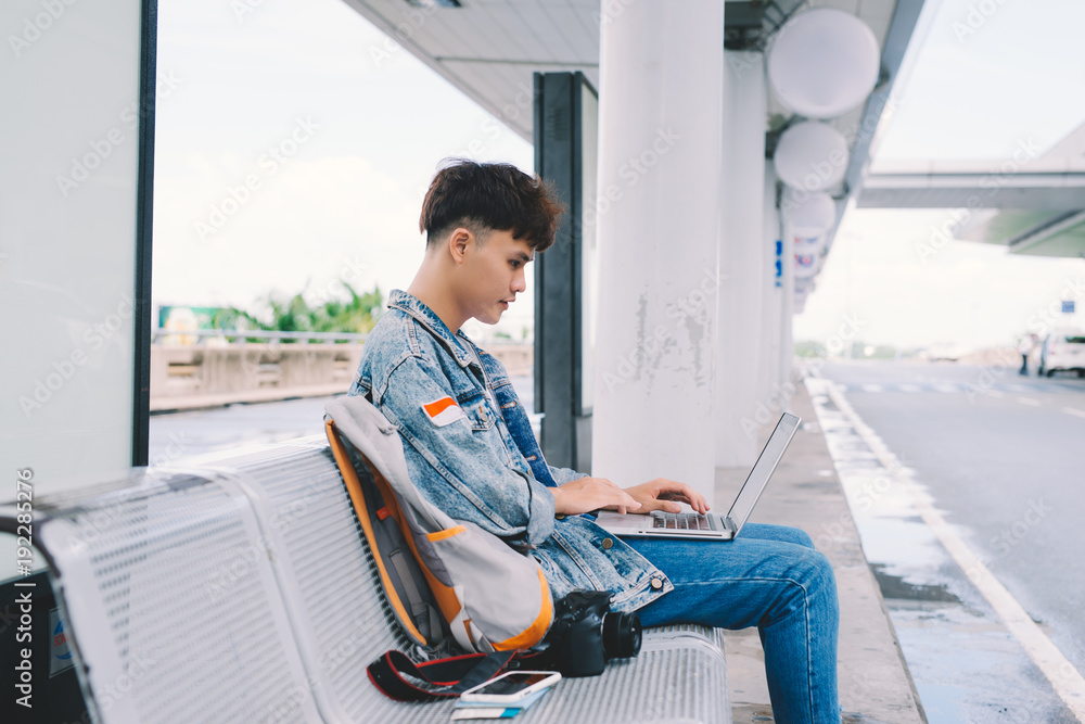 Asian young man sitting on the chair at the airport bus stop and using laptop