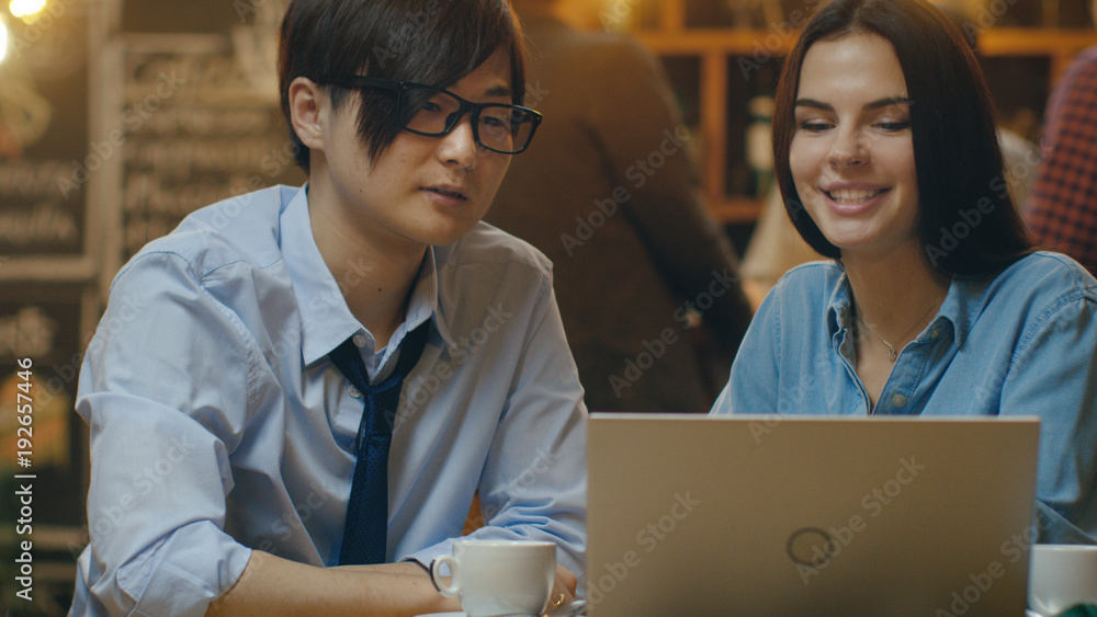 英俊的亚洲男人和美丽的高加索年轻女人坐在咖啡馆里用笔记本电脑工作