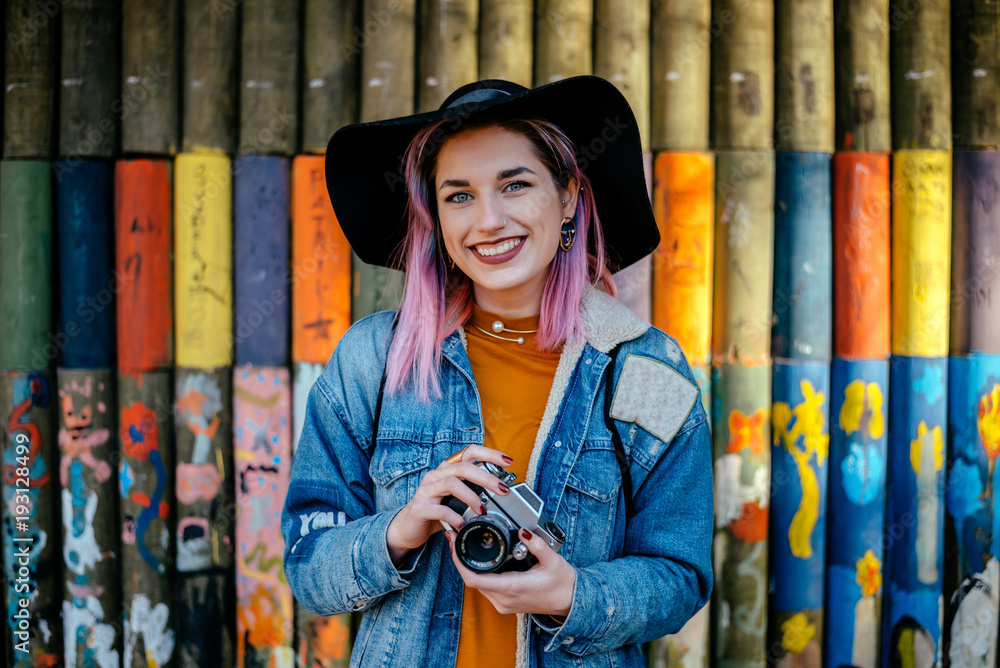 Portrait of a tourist girl with dyed hair and a hat standing in front of a painted wall.