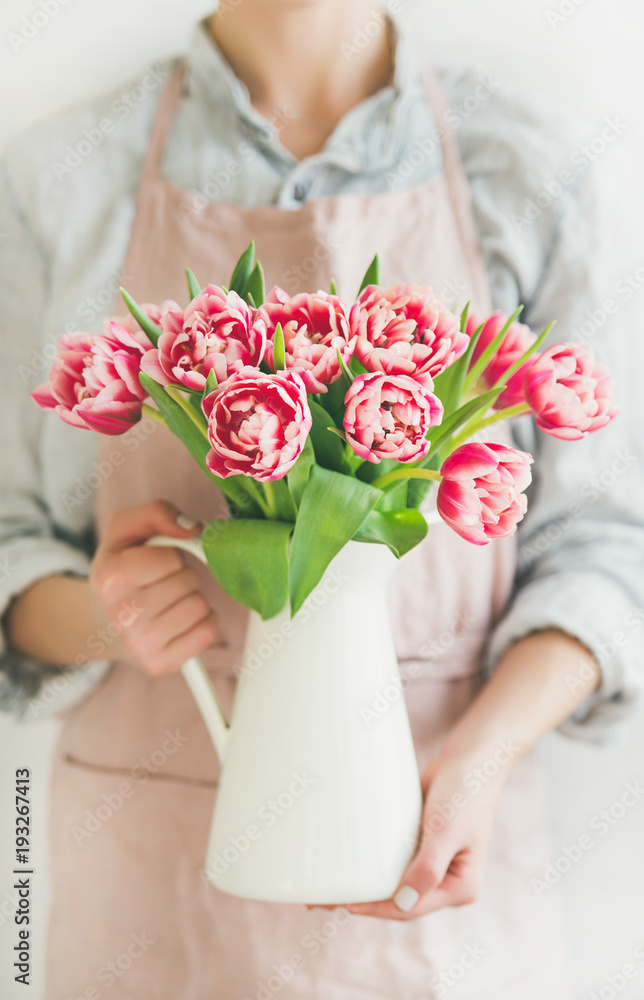 Spring flowers arrangement. Woman in pastel pink apron and blue linen shirt holding white enamel vas