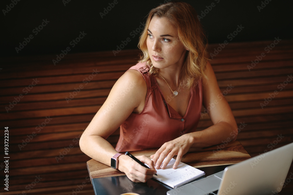 Woman writing notes sitting at a coffee shop with a laptop on the table