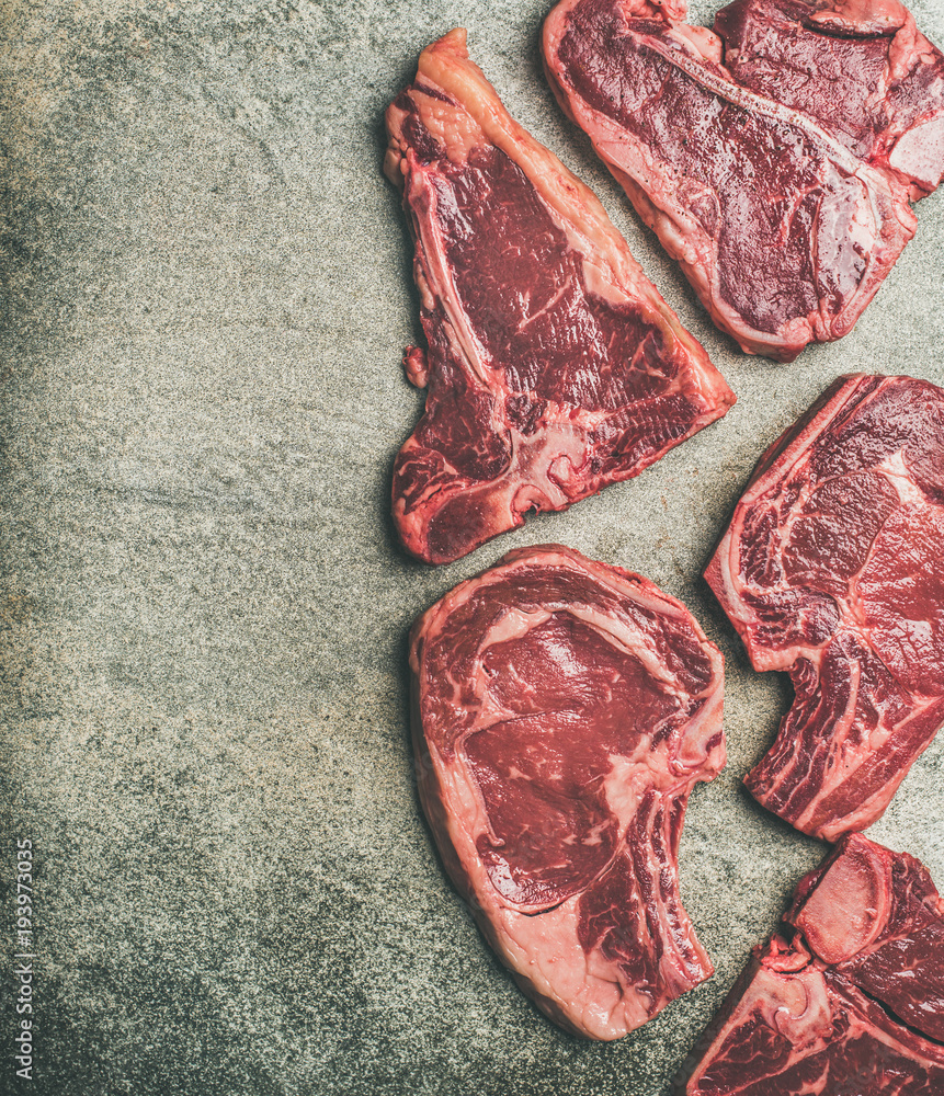 Flat-lay of fresh raw beef meat steak cuts over grey concrete background, top view, copy space, vert