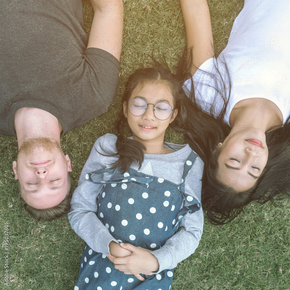 Young family with daughter laying in the park on the green grass and looking at the camera, top view