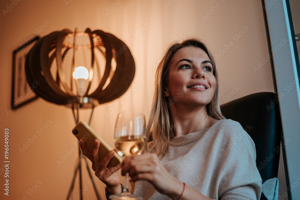 Close-up image of smiling woman enjoying glass of white wine at evening. Using smartphone.