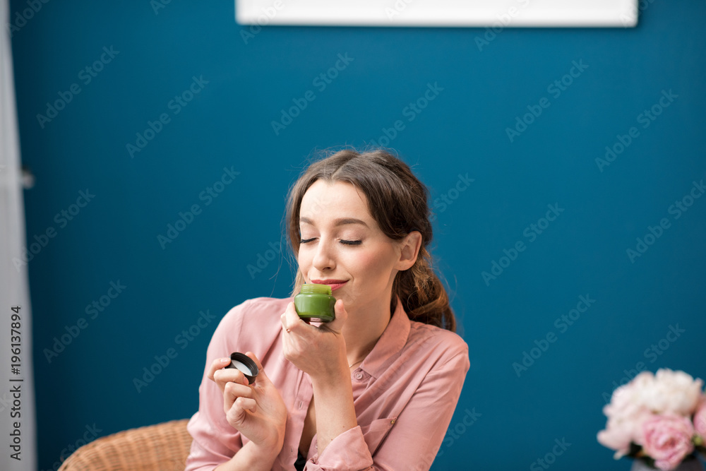 Young woman enjoying aroma of a body lotion sitting on the blue wall background at home