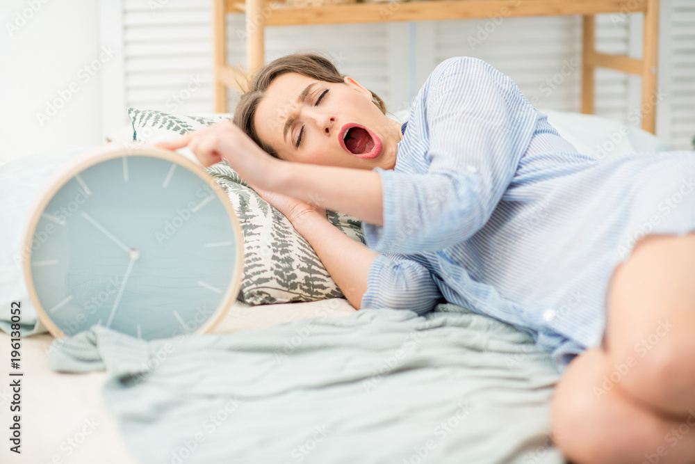 Young woman turning off a big alarm clock trying to wake up yawning on the bed at home