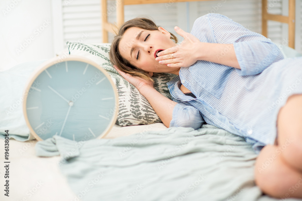 Young and beautiful woman in blue shirt sleeping with big alarm clock on the comfortable bed