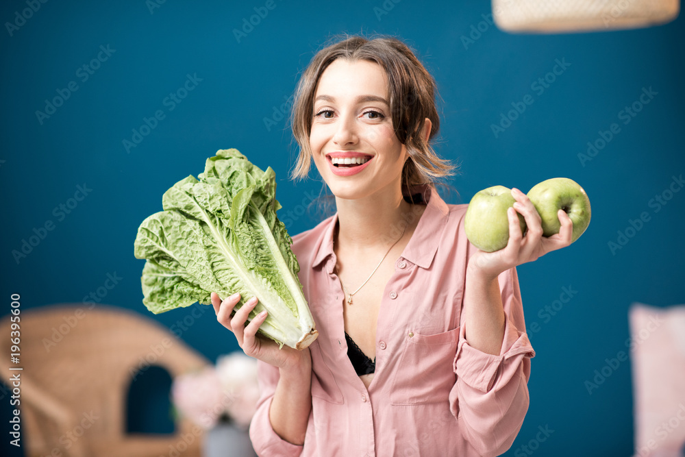 Portrait of a young woman with green lettuce and apples on the blue wall background