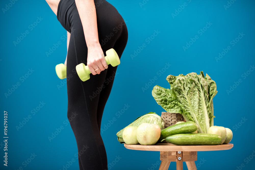 Sports female body with dumbbells and healthy green food on the blue wall background