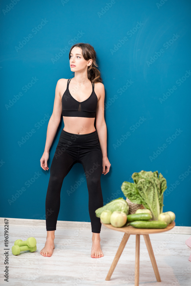 Young woman exercising indoors with green healthy food on the table on the blue wall background at h