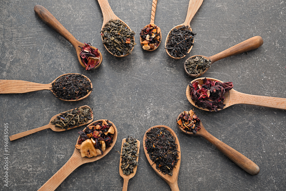 Spoons with different types of dry tea leaves on grey background